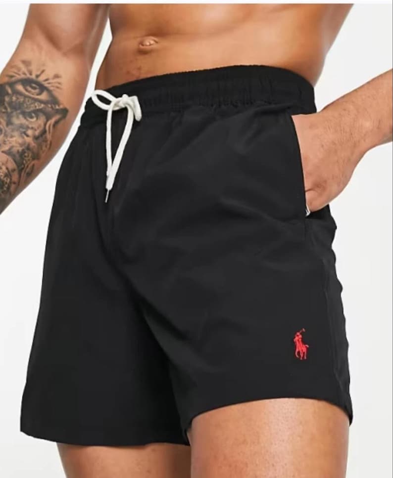 May include: Black swim shorts with a drawstring waist and a red logo on the leg. The shorts have a relaxed fit and pockets. Perfect for a day at the beach or pool.