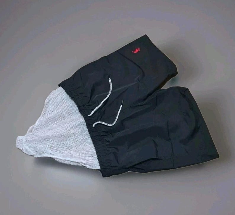 May include: Black swim trunks with a white mesh lining. The trunks have a drawstring closure with white cords and a small red logo on the upper left side. The shorts are made of a lightweight, water-resistant material.