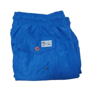 May include: Bright blue swim trunks with a small pocket on the back. The pocket features a label with a palm tree and text. The trunks have a drawstring waistband and a button closure. The fabric has a slight sheen.