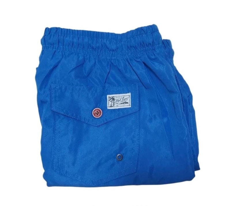 May include: Bright blue swim trunks with a small pocket on the back. The pocket features a label with a palm tree and text. The trunks have a drawstring waistband and a button closure. The fabric has a slight sheen.