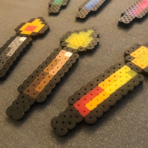 May include: A set of five colorful pixel art wands with black outlines. The wands are made of small plastic beads and feature different color combinations, including red, yellow, orange, brown, and gray.