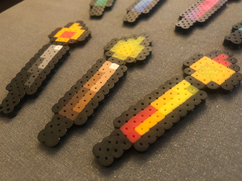 May include: A set of five colorful pixel art wands with black outlines. The wands are made of small plastic beads and feature different color combinations, including red, yellow, orange, brown, and gray.