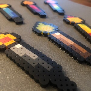 May include: A collection of colorful pixel art wands made with perler beads. The wands are in various colors, including red, yellow, blue, green, and brown.