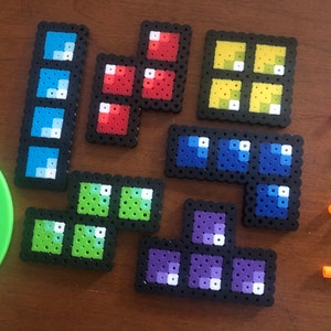 May include: A set of eight colorful pixel art pieces, each depicting a different shape or design. The pieces are made of small, square beads and are arranged on a wooden surface. The colors include blue, red, yellow, green, and purple.