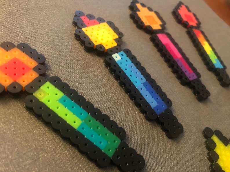 May include: Five colorful pixel art wands made with perler beads. The wands are in a rainbow color scheme, with each wand featuring a different color combination. The wands are all about the same size and are arranged in a row.