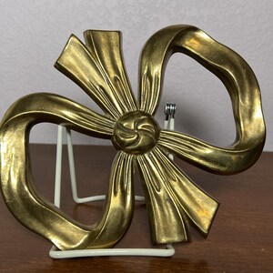 May include: A gold-colored metal bow sculpture with a detailed center knot and flowing ribbon loops. The sculpture is displayed on a white wire stand, set against a neutral background. The bow's metallic finish reflects light, highlighting its intricate design.