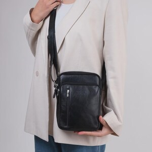 Könnte beinhalten: Eine schwarze Leder-Umhängetasche mit rechteckiger Form und einer Reißverschlusstasche vorne. Die Tasche hat einen langen, verstellbaren Riemen und einen silberfarbenen Reißverschluss. Die Tasche wird von einer Person getragen, die einen cremefarbenen Blazer trägt.