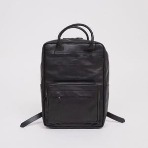 Puede incluir: Mochila de cuero negro con asa superior y correas ajustables. La mochila tiene un bolsillo frontal con cremallera y un pequeño logotipo en relieve en la parte delantera. La bolsa tiene forma rectangular y un diseño clásico.