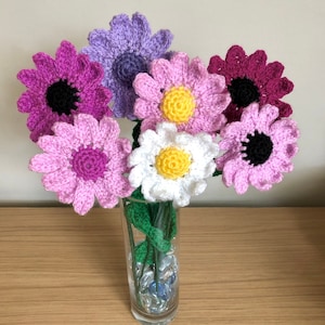 May include: A bouquet of crocheted flowers in a glass vase. The flowers are pink, purple, and white. The vase is filled with clear glass stones.