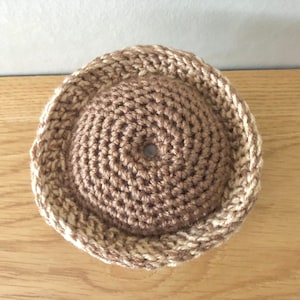 May include: A crocheted hat in brown and beige tones. The hat is round and has a textured surface.