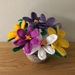 May include: A crocheted bouquet of purple, yellow and white flowers in a brown pot. The flowers are arranged in a vase and have green stems.
