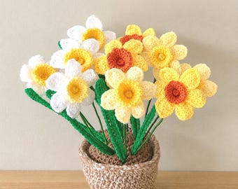 May include: A crochet flower arrangement featuring white and yellow daffodils with orange centres. The flowers have green stems and are planted in a textured, light brown crocheted pot. The petals are crafted with visible yarn stitches.