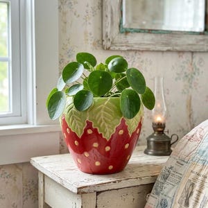 May include: A red and green ceramic strawberry-shaped plant pot with yellow dots, filled with a green plant. The pot sits on a white, distressed wooden table. A vintage oil lamp and a mirror are in the background, creating a rustic aesthetic.