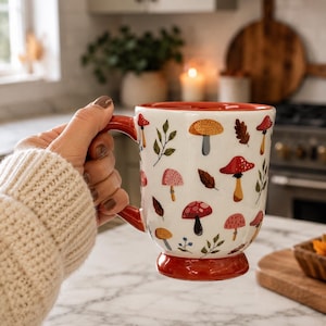 May include: A white ceramic mug featuring a colorful pattern of various mushrooms, leaves, and small branches. The mug has a red handle, a red interior rim, and a red pedestal base. The exterior surface is decorated with red, orange, and brown illustrations.