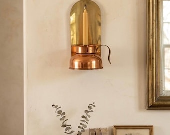 May include: A wall-mounted candle sconce featuring a polished brass backplate and a copper-toned metal cup base. A single cream-colored taper candle is inserted into the center of the cup. The sconce is mounted on a plain, light-colored wall.