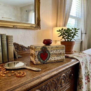 May include: A wooden dresser with a decorative mirror, books, and a small box with a red heart design. A hand mirror and a necklace are on the dresser. A small potted plant and an apple are also visible.