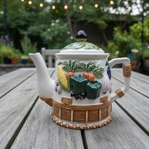 Peut inclure: Théière en céramique blanche avec couvercle et anse. La théière est décorée de légumes colorés, dont du maïs, des poivrons et des carottes. La poignée est enveloppée d'un matériau tissé. La théière est posée sur une table en bois.
