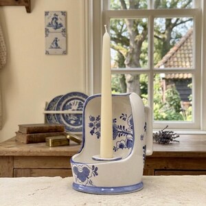 May include: A white ceramic candle holder with blue floral patterns and a handle on the side. The holder features a central circular opening for a single taper candle, which is currently holding a tall, slender, cream-coloured wax candle.