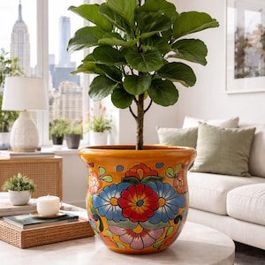 May include: A large, leafy green plant in a vibrant orange ceramic pot with a floral design. The pot sits on a round, marble-topped coffee table. The background includes a window with a city view and a white sofa.