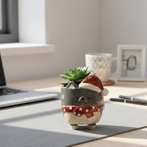 Puede incluir: Una maceta de cerámica gris con forma de gato con un gorro y bufanda de Papá Noel rojos y blancos, que contiene una suculenta verde. La maceta está sobre un escritorio con un portátil, una taza y un marco de fotos.