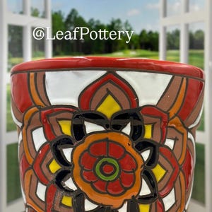 May include: A white ceramic planter with a red rim and a colorful mandala design featuring red, orange, yellow, green, and black. The planter is decorated with a geometric pattern and a floral motif.