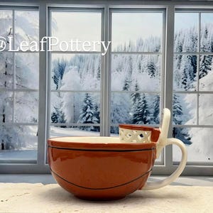 Könnte beinhalten: Keramikbecher im Basketball-Design mit orange-schwarzem Farbschema. Der Becher hat einen Griff und ein kleines korbförmiges Detail. Der Hintergrund zeigt eine verschneite Winterlandschaft.