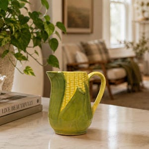 May include: A ceramic pitcher shaped like a corn cob with a yellow kernel body and green husk leaves forming the base and handle. The pitcher features a curved spout and a sturdy handle, showcasing a textured, realistic design of a corn cob.