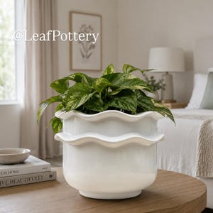 May include: A white ceramic planter with a double-tiered wavy rim containing a green and yellow variegated pothos plant. The text @LeafPottery is visible in the upper left corner of the image, positioned above the plant.