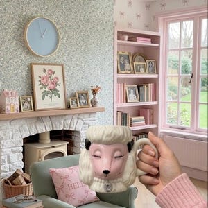 以下が含まれることがあります： 居心地の良いリビングルームのシーンの前に、目を閉じたプードルの形をしたセラミックマグカップ。 マグカップはクリーム色で、黒のアクセントとシルバーのチャームが付いています。 部屋には暖炉、緑のアームチェア、ピンクのサイドテーブルがあります。