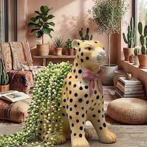 May include: A ceramic leopard figurine planter with a glossy cream finish and black spots. The leopard features a sculpted pink bow around its neck and contains a trailing string of pearls plant cascading from its back, creating a decorative display.
