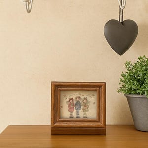 Puede incluir: Una imagen enmarcada con un marco de madera que muestra tres figuras con vestidos. Un adorno en forma de corazón gris oscuro cuelga de una estantería decorativa de metal. Una pequeña planta en maceta se encuentra sobre una superficie de madera.