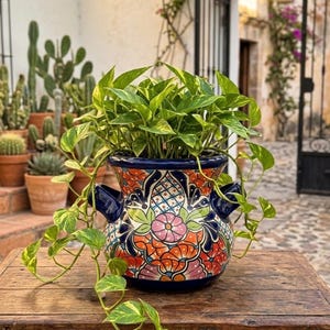Może przedstawiać: Żywa, ręcznie malowana ceramiczna donica z kolorowym kwiatowym wzorem w kolorach pomarańczowym, różowym i niebieskim. Donica zawiera bujną zieloną roślinę z pnączami. Donica stoi na rustykalnym drewnianym stole, tworząc uroczy, dekoracyjny wyświetlacz.