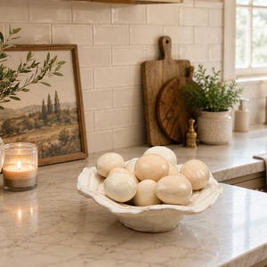 May include: A white, scalloped-edge ceramic bowl filled with a collection of smooth, cream-colored decorative eggs. The bowl has a textured, organic shape and sits on a marble countertop, showcasing a glossy finish on the rounded egg surfaces.