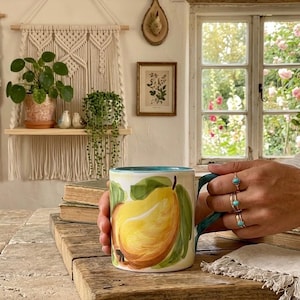 May include: A ceramic mug with a hand-painted yellow pear design, green leaves, and a turquoise interior. The mug is held by a person with multiple rings on their fingers. The mug is on a wooden table.