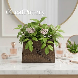 May include: A square, dark brown wooden planter with a textured design, filled with a green plant with pink and white flowers. The planter is on a white marble surface, with a mirror and perfume bottles in the background.