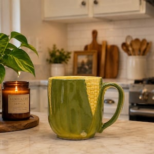 May include: A ceramic mug shaped like a corn cob, featuring a bright yellow textured kernel pattern on the upper portion and smooth green husks covering the lower body and base, complete with a curved green handle on the right side.