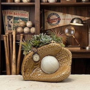 May include: A baseball-themed planter featuring a brown ceramic baseball glove with a white baseball and a watch attached. The glove is filled with succulents and greenery. The background includes baseball bats, vintage baseballs, and a pennant.