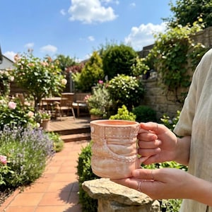 May include: A peach-colored ceramic mug with a textured design, featuring a handle and a detailed pattern. The mug is held in front of a garden scene with lush greenery, flowers, and a stone wall, under a bright blue sky.