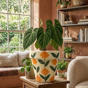 May include: A large, cream-colored ceramic plant pot featuring a hand-painted pattern of orange flowers with dark green leaves and stems. The pot contains a large green Anthurium plant with prominent heart-shaped leaves and a single white spathe.
