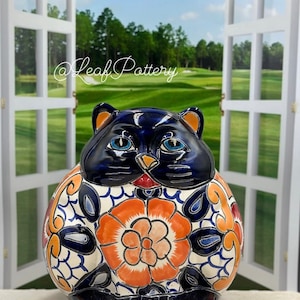 May include: A hand-painted ceramic cat figurine with blue and orange floral designs. The cat is sitting with its paws tucked underneath it. The figurine is a traditional Mexican folk art style.
