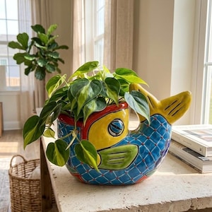 May include: A ceramic fish-shaped planter featuring a vibrant blue body with a red grid pattern, a yellow tail and face, and green accents. The planter holds a trailing philodendron plant with variegated green and yellow heart-shaped leaves.
