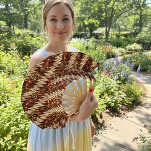 Puede incluir: Abanico de mano decorativo con un diseño estampado marrón y beige. El abanico tiene un marco de madera y se sostiene abierto. La tela del abanico presenta un patrón repetido de formas florales y geométricas. Un accesorio elegante.