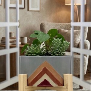 May include: A rectangular gray planter box with a geometric design in shades of brown and white, holding green succulents and a plant with large, round leaves. The planter sits on a wooden stand with a decorative front.