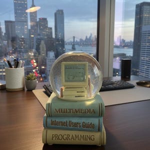 May include: A snow globe featuring a vintage computer model, resting atop a stack of books with titles like "Multimedia" and "Internet Users Guide." The globe sits on a desk with a city skyline visible through a window.