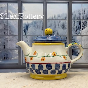 Op de afbeelding: Een keramische theepot met een blauw deksel en gele knop. De body is crèmekleurig met een bloemenmotief en een blauw-wit geblokt patroon aan de basis. De handgreep en tuit zijn wit.