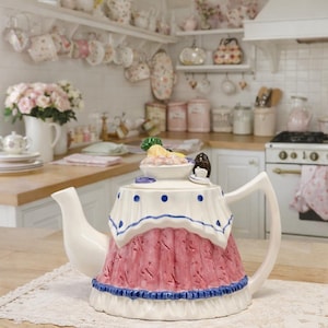 Puede incluir: Una tetera de cerámica decorativa con base blanca, diseño de falda rosa y ribete azul. La tapa está adornada con un plato pequeño con comida y una pequeña figura. La tetera está colocada sobre un mantel de encaje en un entorno de cocina.