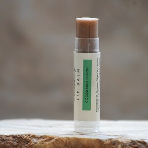 Könnte beinhalten: Ein weißer Lippenbalsam-Stift mit einem grünen Etikett, auf dem "Cocoa Mint Fusion" steht und die Zutaten aufgelistet sind: Bio-Olivenöl, Kokosöl, Rizinusöl, Bienenwachs, Bio-Kakao.