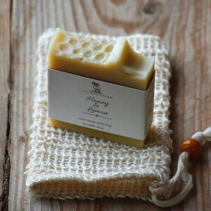 Puede incluir: Una barra de jabón artesanal en una bolsa de malla blanca. El jabón es de color amarillo claro y tiene un patrón de panal. La etiqueta del jabón dice "N.A. Wild Touch Honey & Lemon 100% Handcrafted Soap Net Weight 120 gr."