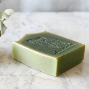 Puede incluir: Una pastilla de jabón verde con un diseño en relieve de una persona sosteniendo una planta. El jabón está sobre una superficie de mármol blanco.