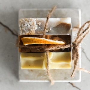 Puede incluir: Cuatro barras de jabón envueltas en cuerda y apiladas sobre una superficie de mármol blanco. Las barras de jabón son de diferentes colores y tienen un aspecto natural y hecho a mano.